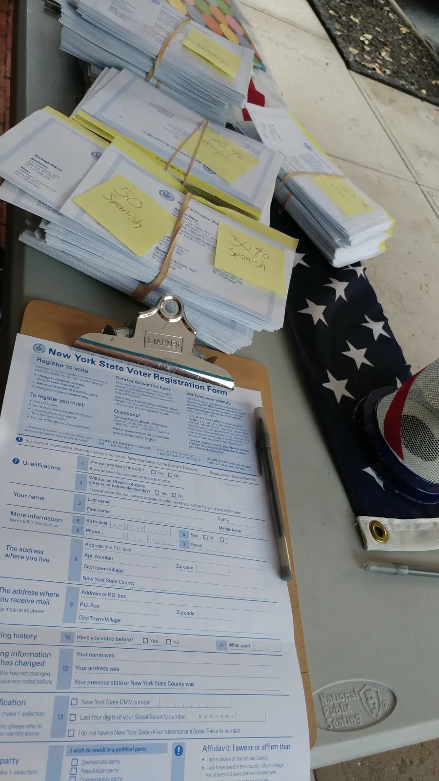 Formularios de registro de votantes de Nueva York sobre una tabla con sujetapapeles, junto a una bandera estadounidense