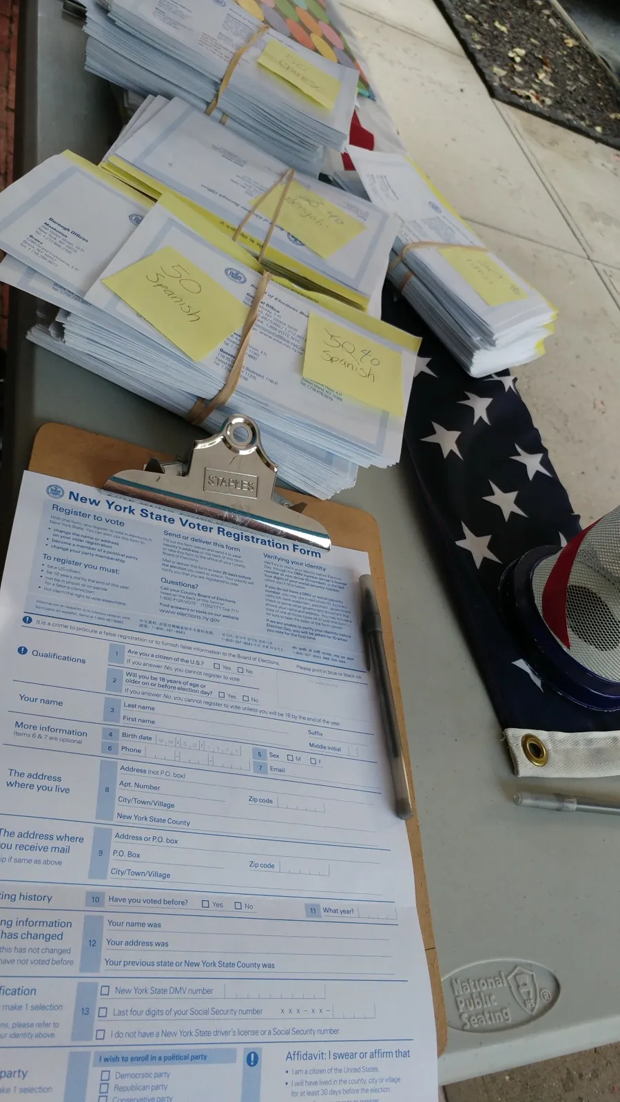 Formularios de registro de votantes de Nueva York sobre una tabla con sujetapapeles, junto a una bandera estadounidense