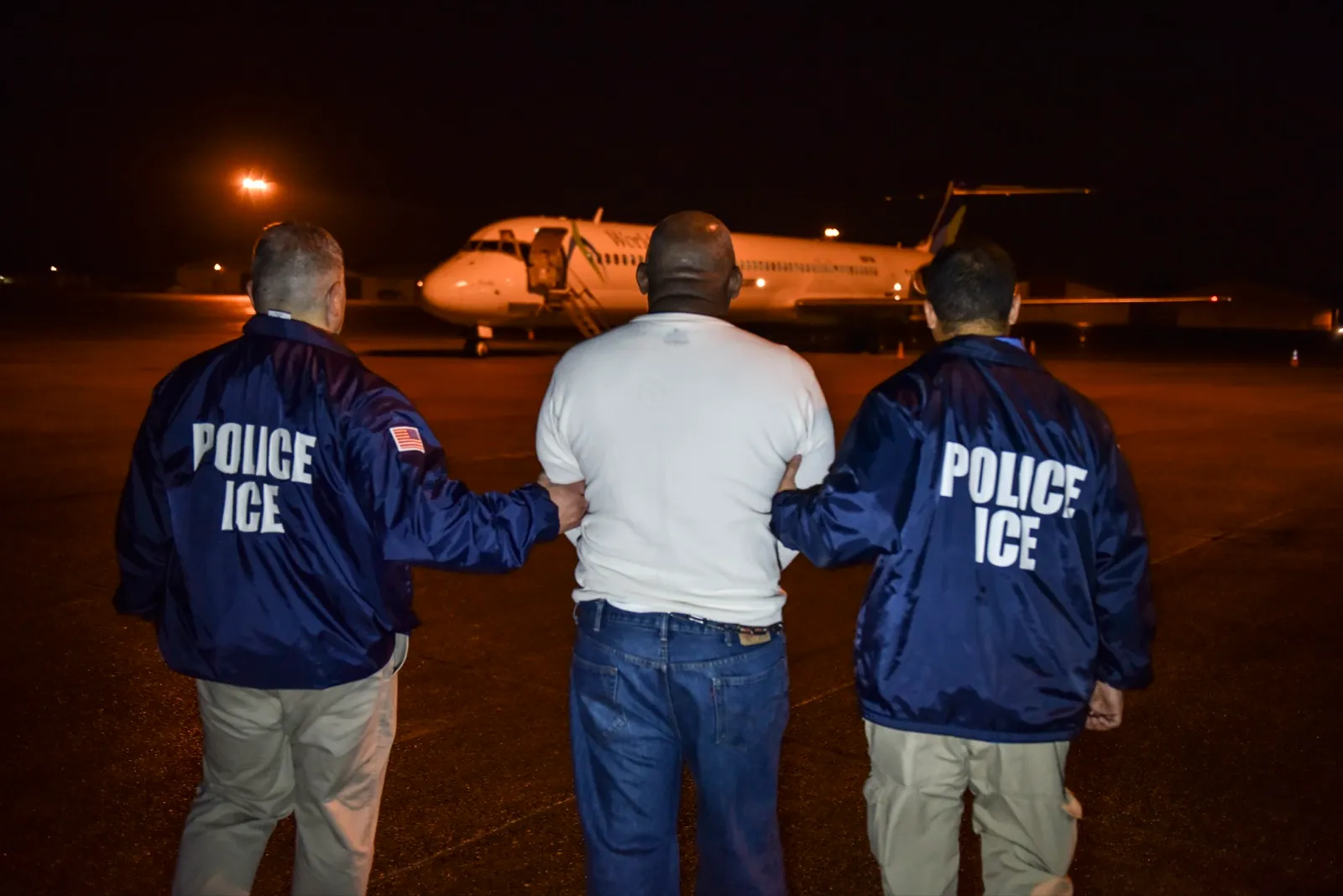 Dos oficiales de ICE escoltan de espaldas a un detenido hacia un avión de vuelos de deportación, de noche