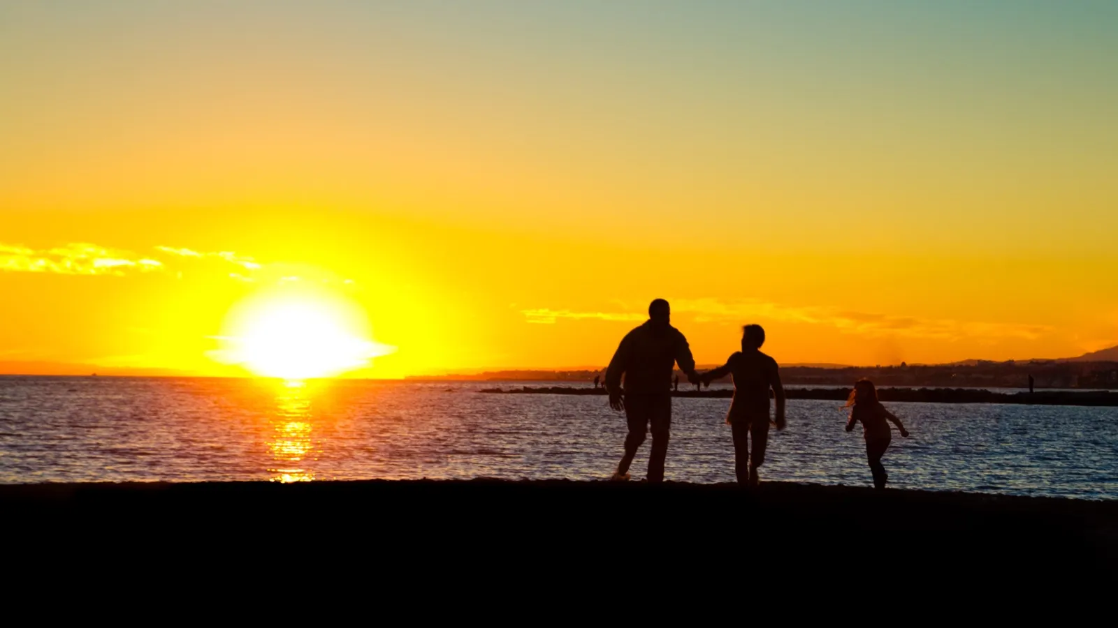 Siluetas de dos adultos y una niña caminando por la playa al atardecer