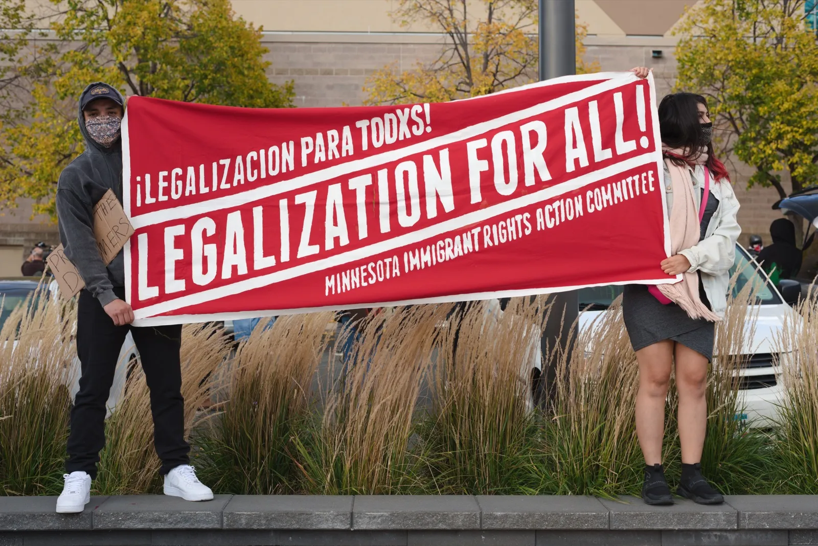 Manifestantes con mascarilla sostienen una pancarta roja que pide legalización para todos durante una marcha por el TPS y DACA